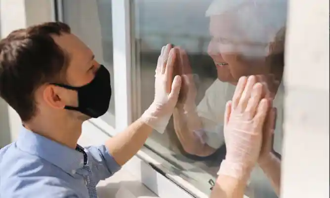 Man in face mask holds his hands up to the window of his Grandma, who holds her hands up to the glass too